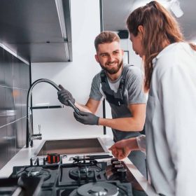 young-professional-plumber-in-grey-uniform-shows-results-of-work-for-housewife-on-the-kitchen